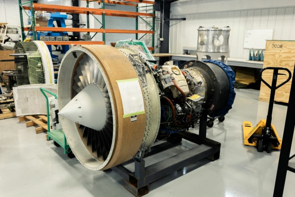 Jet engine assembly in a maintenance facility, featuring a large turbofan with exposed wiring and components, mounted on a stand for inspection or repair. Surrounding the engine are tools, pallets, and industrial shelving in a clean, organized workspace.