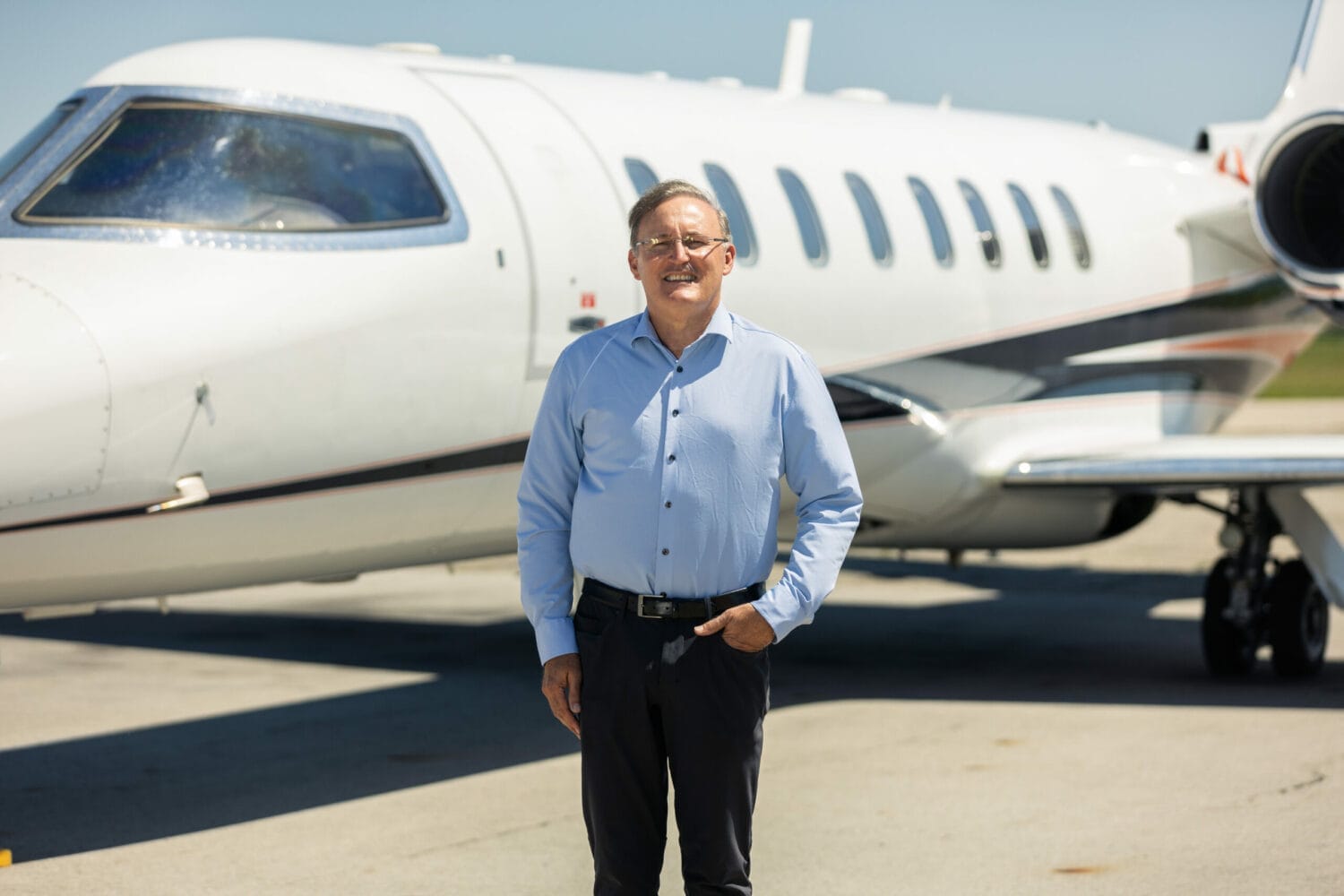 Doug Kalitta standing in front of a private jet on an airport tarmac, wearing a light blue dress shirt and dark trousers.