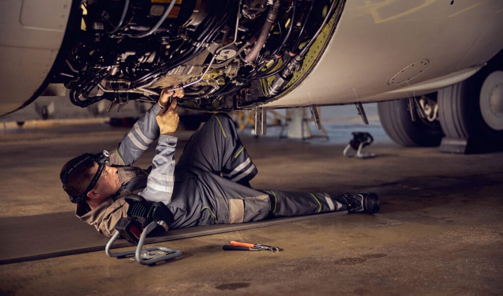 Aircraft maintenance technician working beneath an aircraft in a hangar, performing detailed inspection and engine repairs using reliable replacement parts to maintain optimal safety and performance standards.